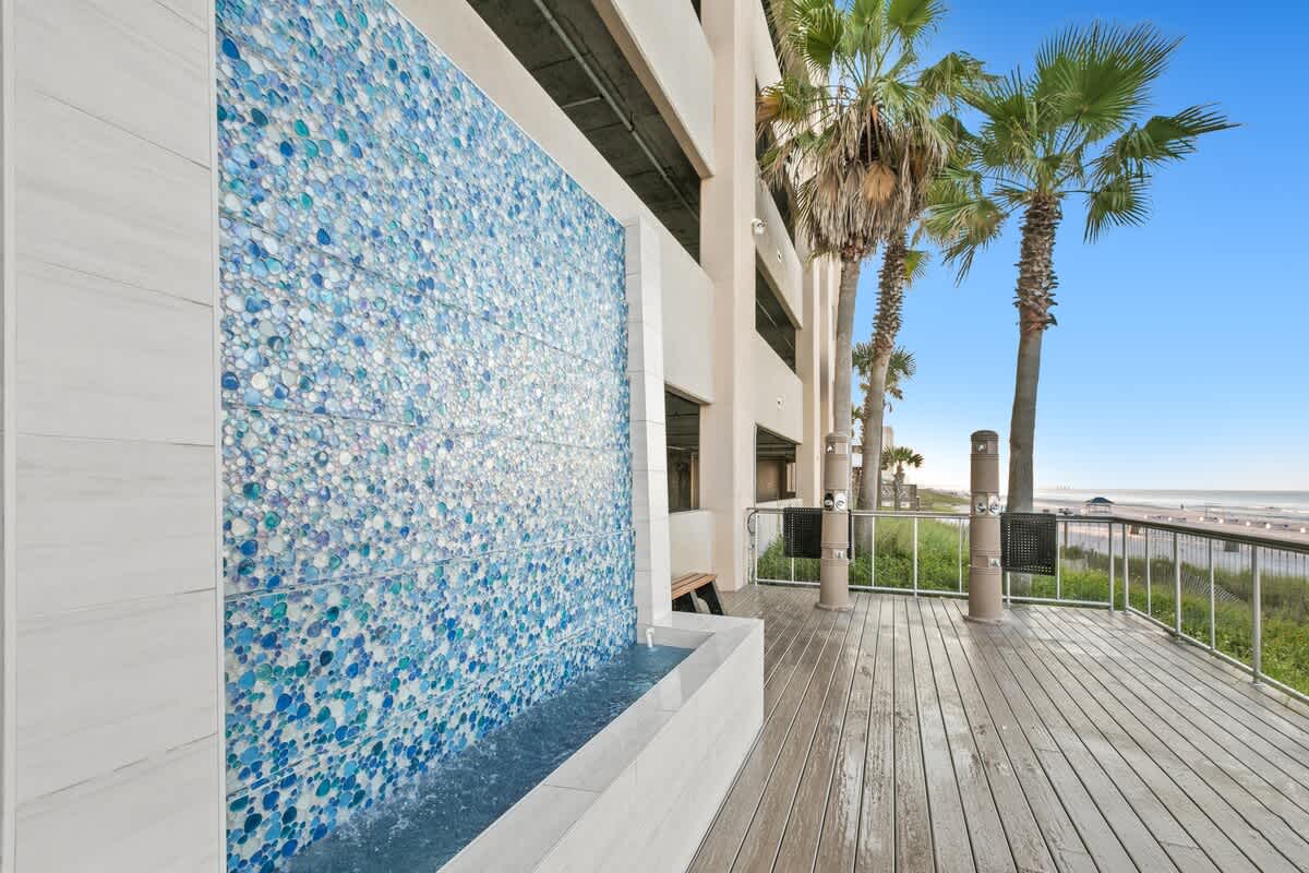 View of an outdoor wooden deck with a striking blue mosaic water wall, offering glimpses of the ocean and sandy beach in the distance. Palm trees line the side of the building, enhancing the tropical feel.