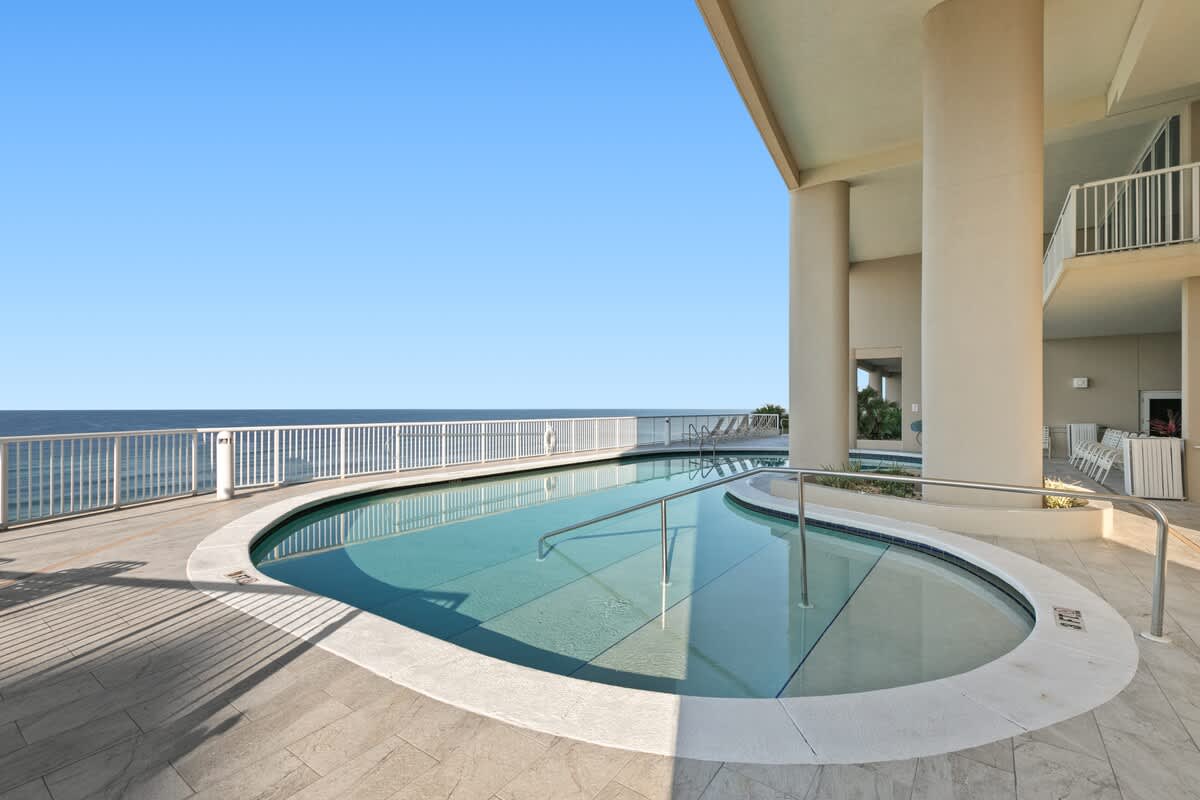A curved outdoor communal pool with a shallow entry is set on light-colored tiled flooring, offering expansive views of the ocean. The pool area is partially shaded by the building's overhang, with a white railing separating it from the sea.