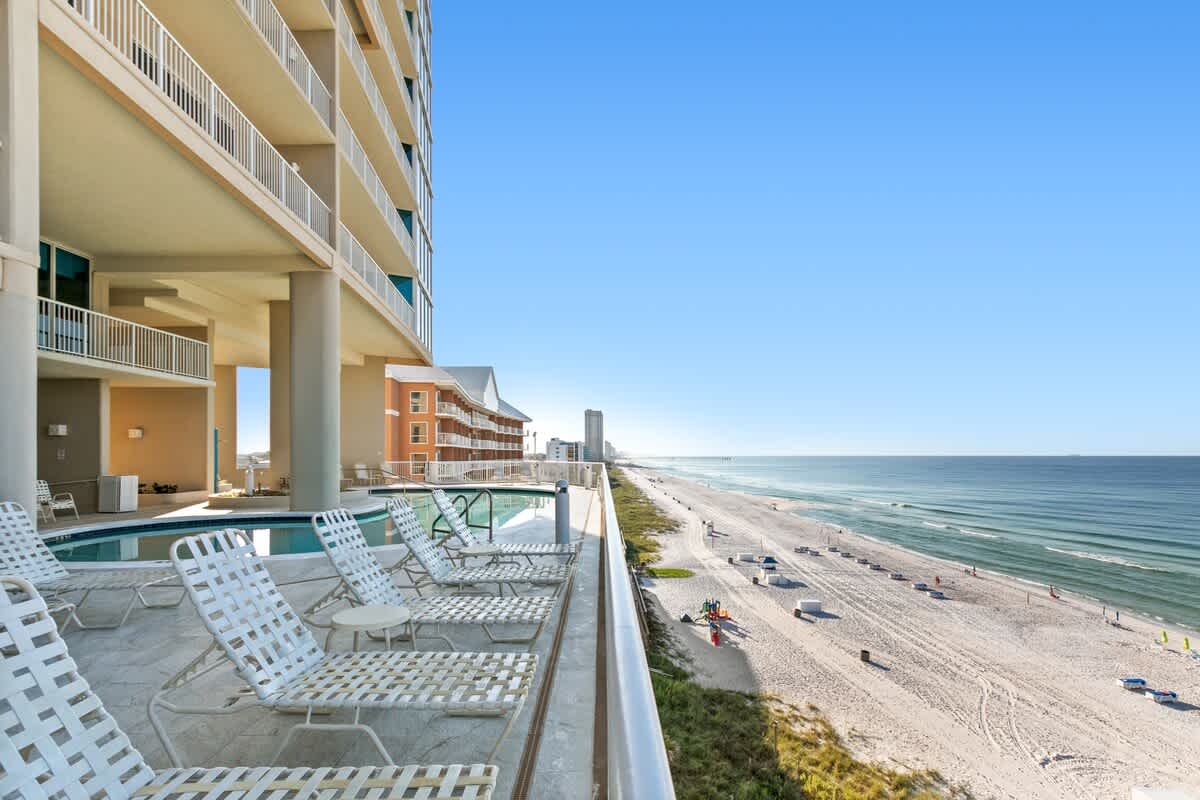 An elevated outdoor communal pool area with white lounge chairs, overlooking a vast sandy beach and the ocean under a clear blue sky. A multi-story condominium building stands to the left, providing shade to part of the pool deck.