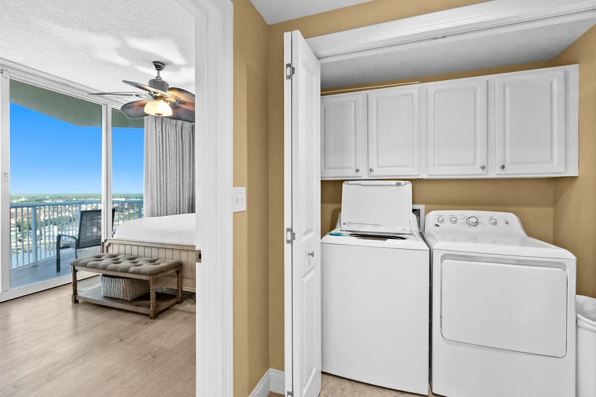 Close-up view of a laundry closet with a white top-loading washer and a white dryer, complemented by white storage cabinets above. The space features light beige walls and a tiled floor, with a bedroom visible through an adjacent doorway.