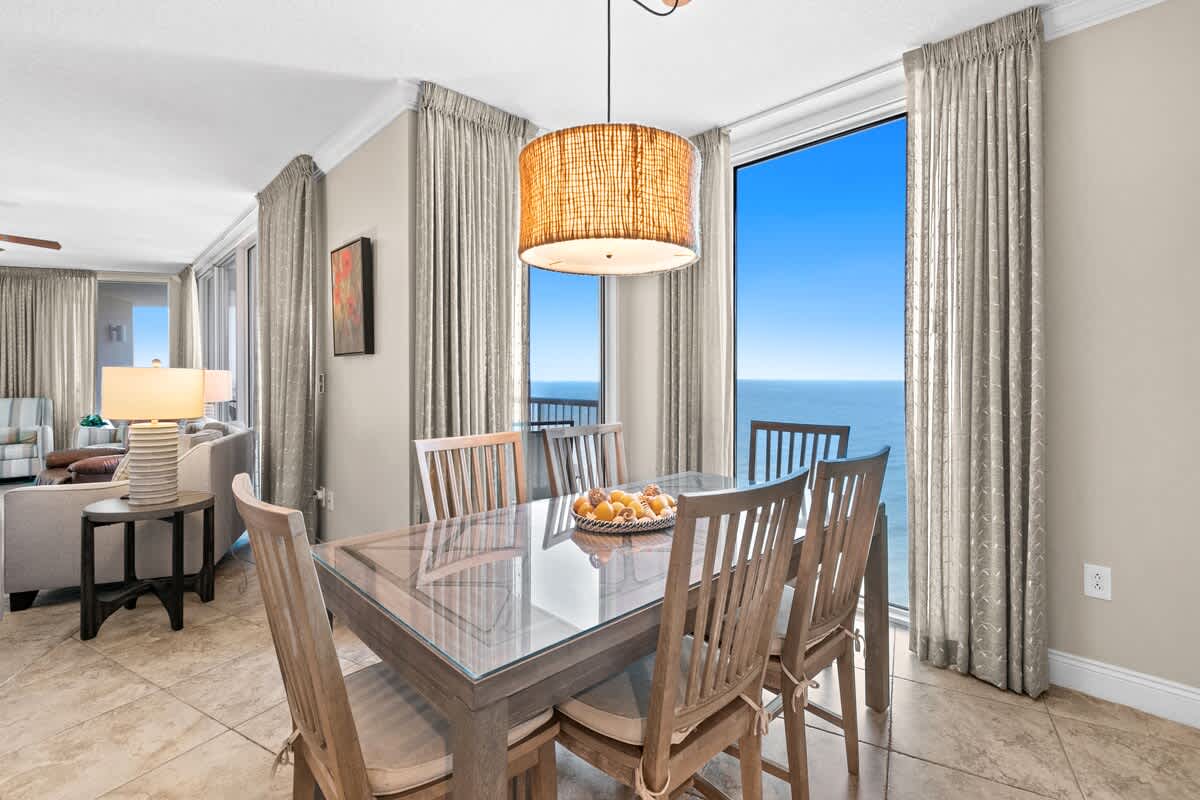 The dining area features a rectangular glass-top table with wooden chairs, illuminated by a woven pendant light, and offers direct ocean views through large windows with light drapes. The room has light tile flooring.