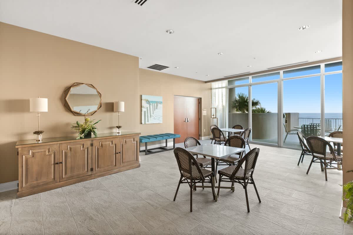 A bright communal lounge area features light beige walls, tiled flooring, and several small square dining tables with dark wicker chairs. A large wooden console table with lamps and a round mirror sits against one wall, and floor-to-ceiling windows offer ocean views.