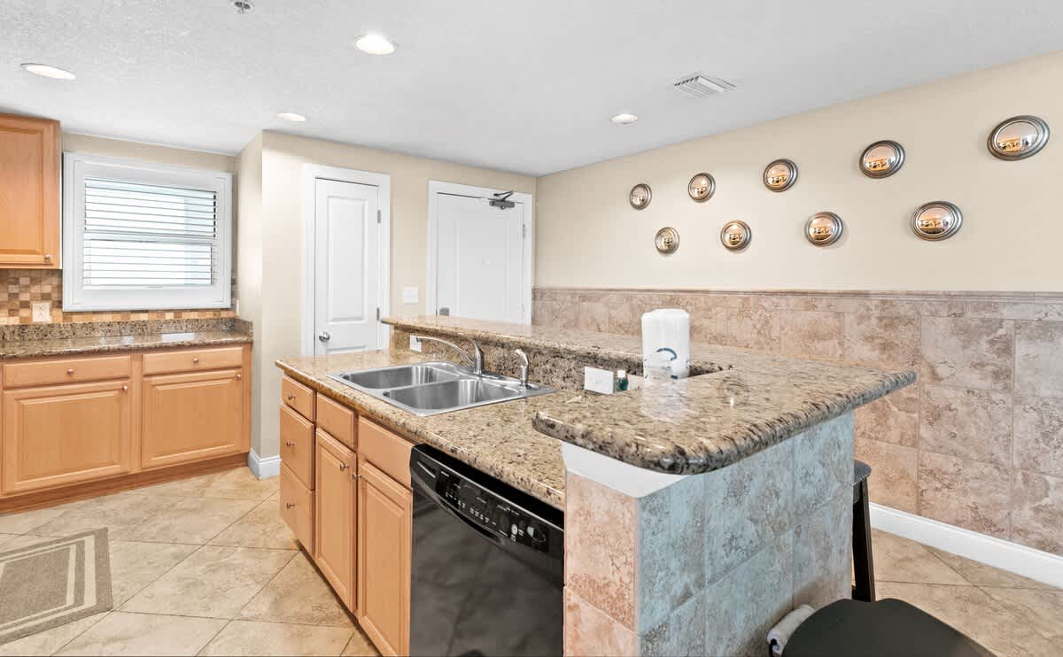 A bright kitchen features light wood cabinetry, granite countertops, and a double stainless steel sink, with a breakfast bar extending into the room and tiled flooring.