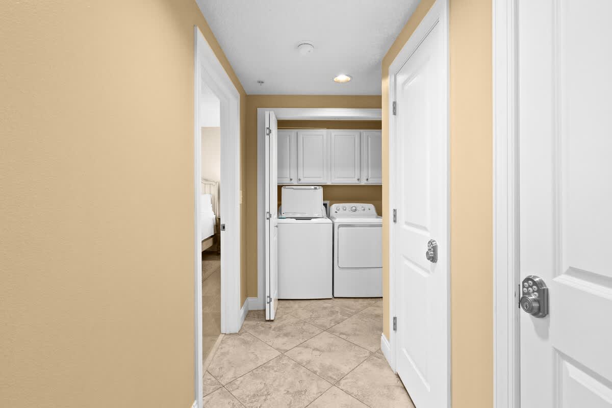 Hallway with light beige walls and tiled flooring, featuring a laundry closet with a white washer and dryer and white upper cabinets. A doorway on the left leads into a bedroom.
