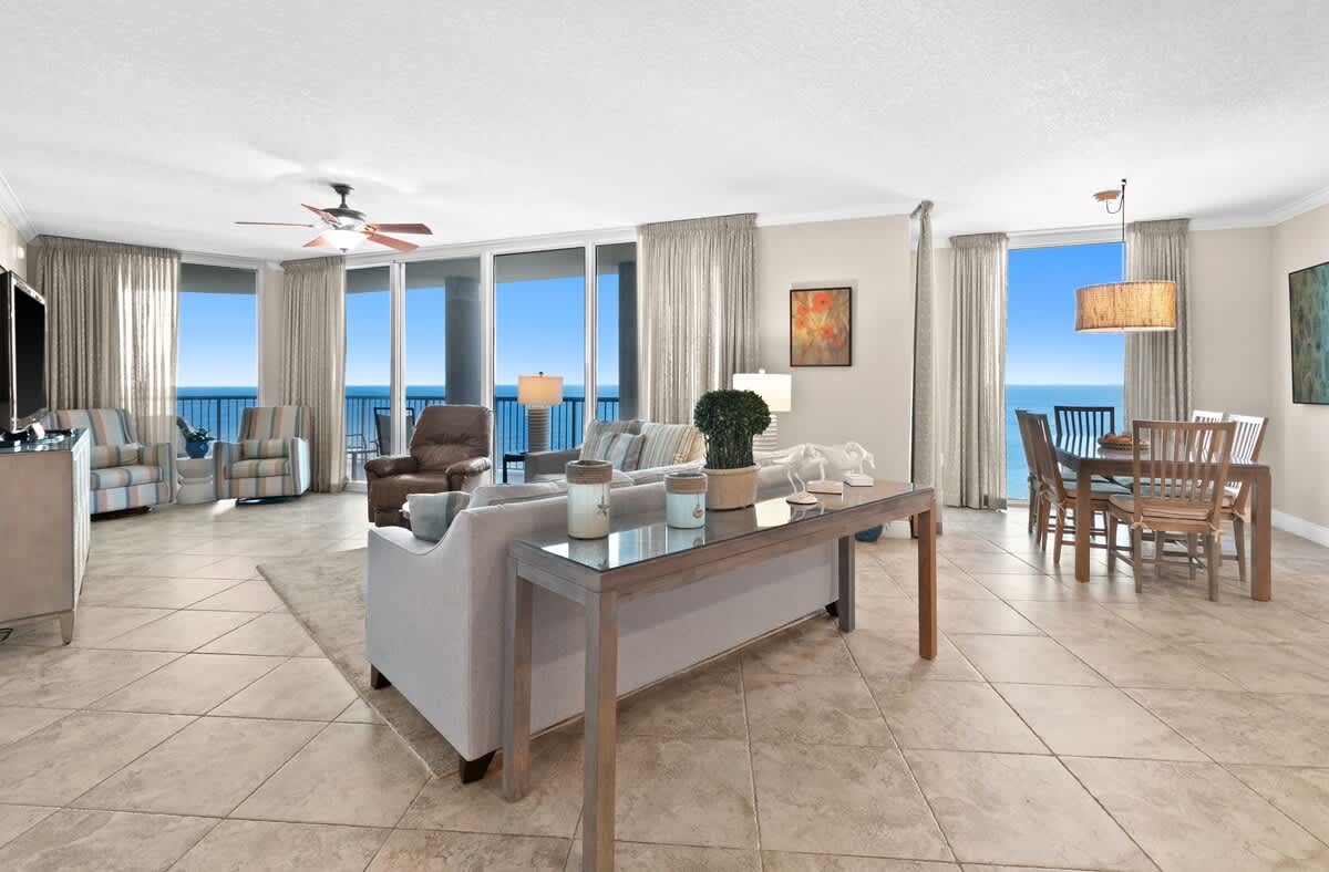 A spacious living room with light-colored sofas and a rug opens to a dining area featuring a glass-top table and wooden chairs, all set against a backdrop of floor-to-ceiling windows with panoramic ocean views. The room has light tile flooring and a ceiling fan.