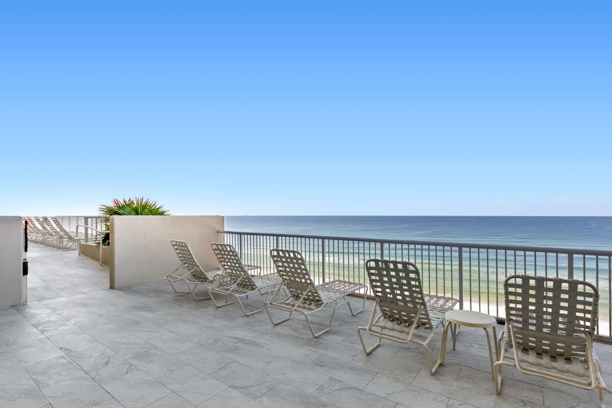 An expansive outdoor deck with light-colored tiled flooring features several white mesh lounge chairs and small side tables. The deck provides an unobstructed, panoramic view of the ocean and coastline, bordered by a white railing.