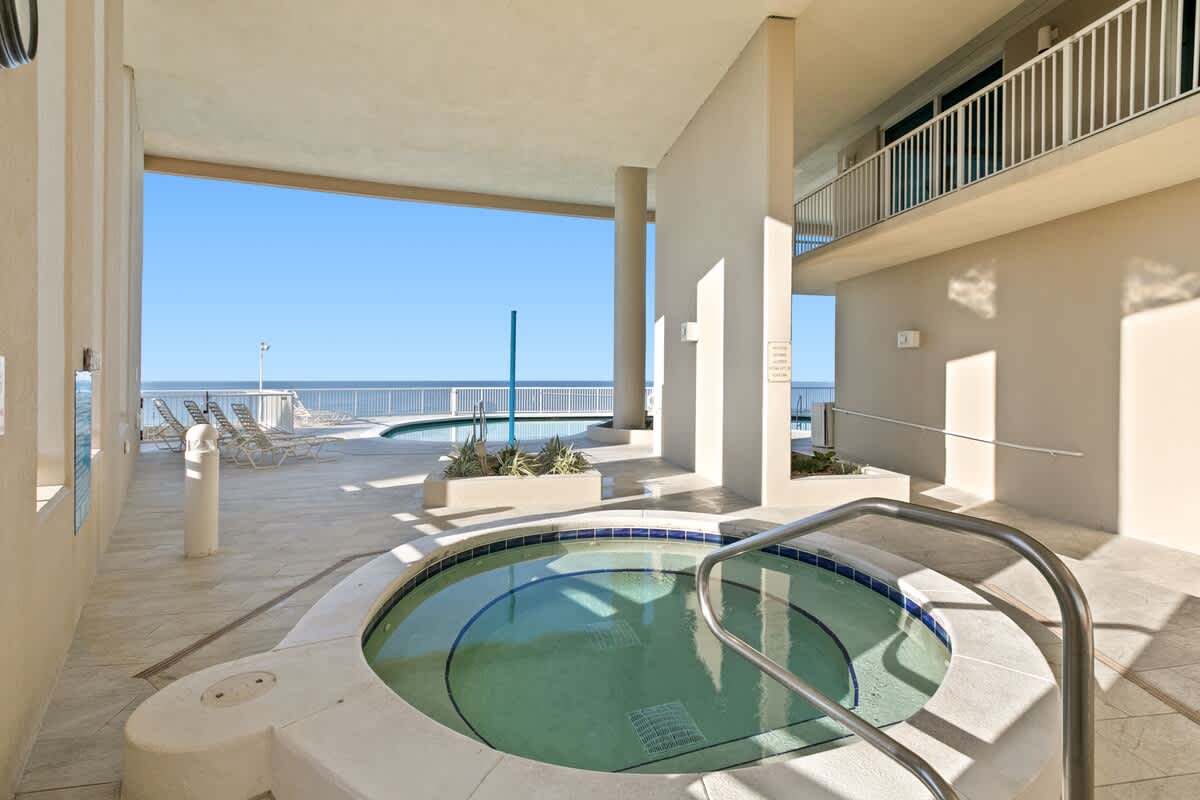 A circular outdoor hot tub with blue tile accents and a metal handrail is set on light-colored tiled flooring, partially shaded by the building's structure. Beyond it, a communal pool and the vast ocean are visible under a clear blue sky.