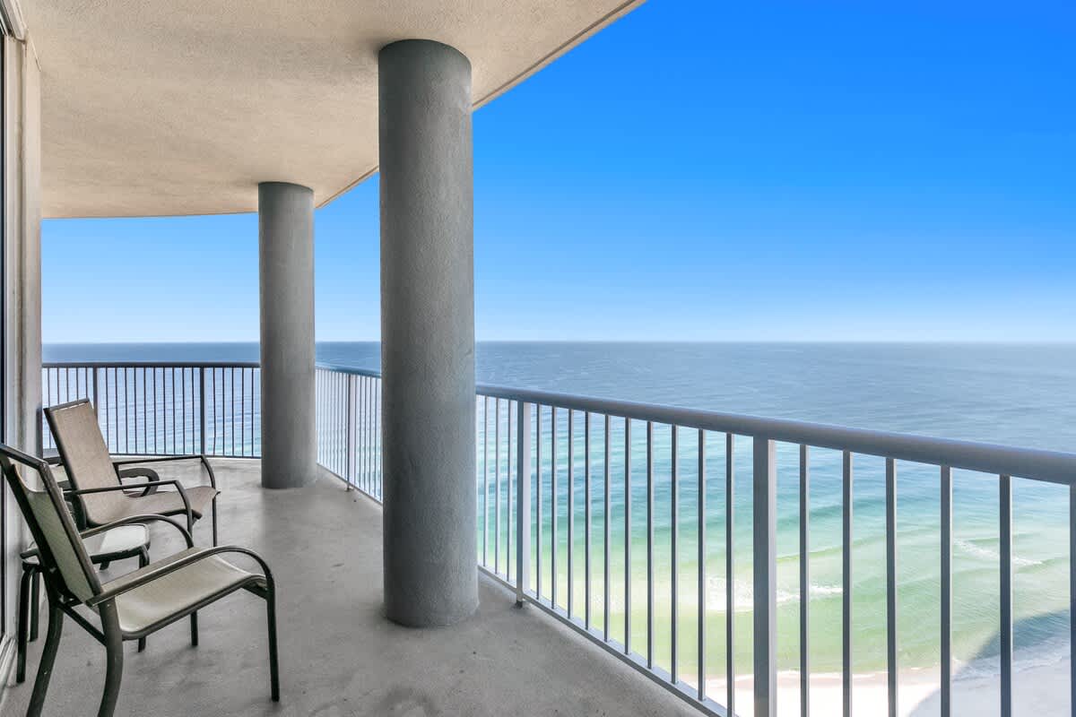 Spacious outdoor balcony with sturdy metal railings and two chairs, offering a breathtaking panoramic view of the vast ocean and distant coastline under a clear blue sky.