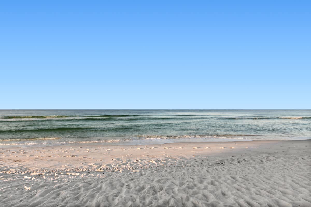 Serene view of the ocean with small waves breaking on a wide, light-colored sandy beach under a vast, clear blue sky. The water displays beautiful shades of blue and green.