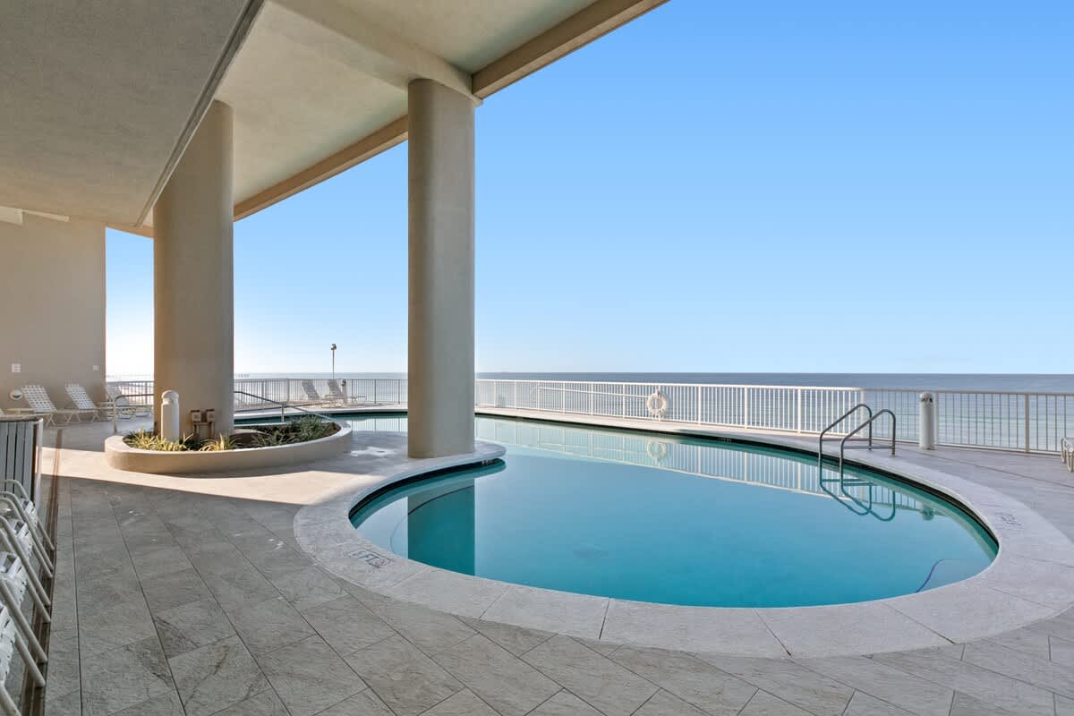 A curved outdoor communal pool with a shallow entry is surrounded by light-colored tiled flooring and partially shaded by the building's structure. The pool offers a direct view of the vast ocean and horizon under a bright blue sky.