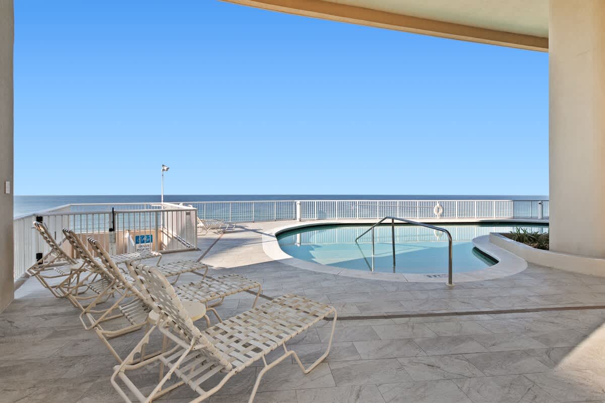 An outdoor communal pool with a shallow entry area is surrounded by light-colored tiled flooring and several white mesh lounge chairs. The pool overlooks the ocean, visible beyond a white railing under a clear blue sky.