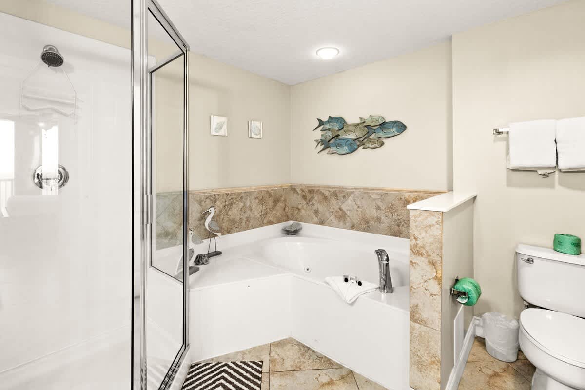 This bathroom includes a corner jetted tub with tiled surround, a toilet, and a glass-enclosed shower, decorated with coastal-themed accents and light-colored tiles.