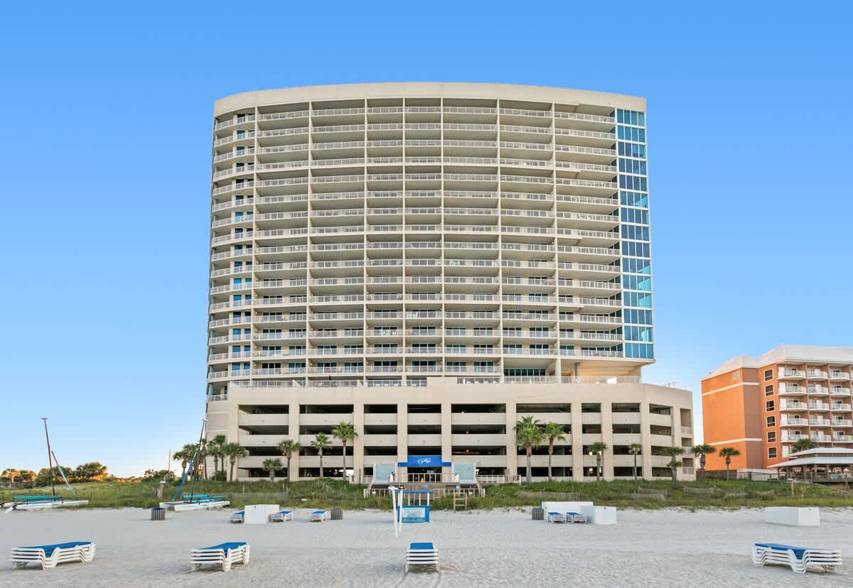Large, modern condominium building with numerous balconies and a multi-level parking garage at its base, viewed from the sandy beach. Lounge chairs are neatly arranged on the sand in front of the property.