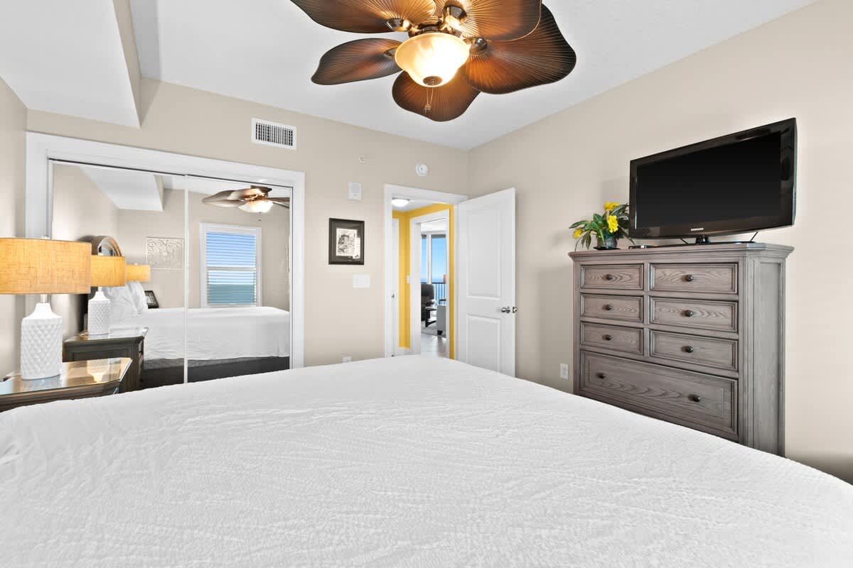 Bright bedroom with light beige walls, a large white bed, a dark wooden dresser with a TV, and a tropical leaf-bladed ceiling fan. A mirrored closet reflects the room, and an open doorway leads to another area.