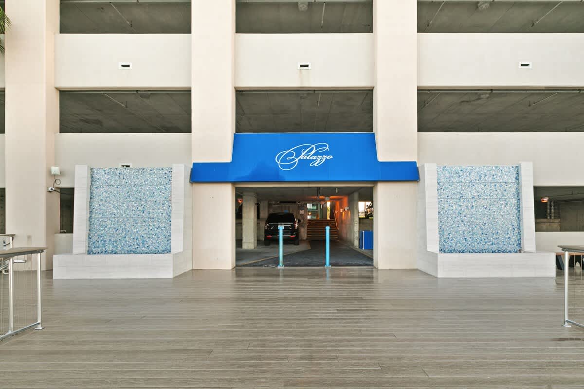The main entrance to the building, featuring a prominent blue awning with 'Palazzo' script above a drive-through area. Two decorative water walls with blue mosaic tiles flank the entrance, adding a touch of elegance.
