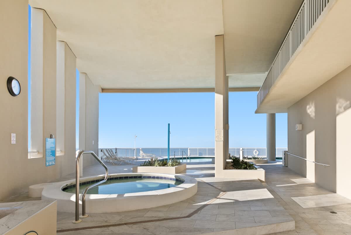 An outdoor hot tub with blue tile accents is surrounded by light-colored tiled flooring and covered by an overhead structure. The ocean is visible in the background, beyond a communal pool and lounge chairs, under a clear blue sky.