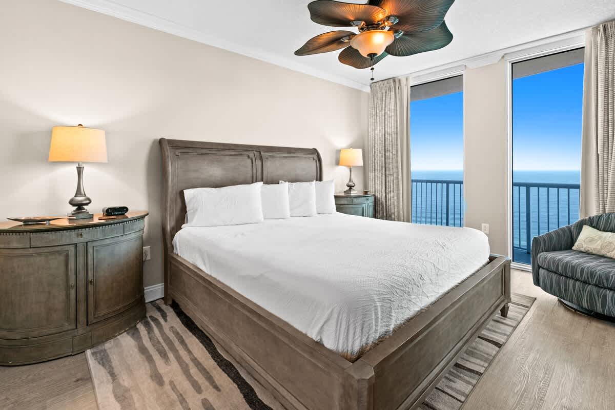 This bedroom features a large king-sized bed with a tall headboard, light wood flooring, and expansive windows showcasing ocean views, with a patterned rug and two nightstands with lamps.