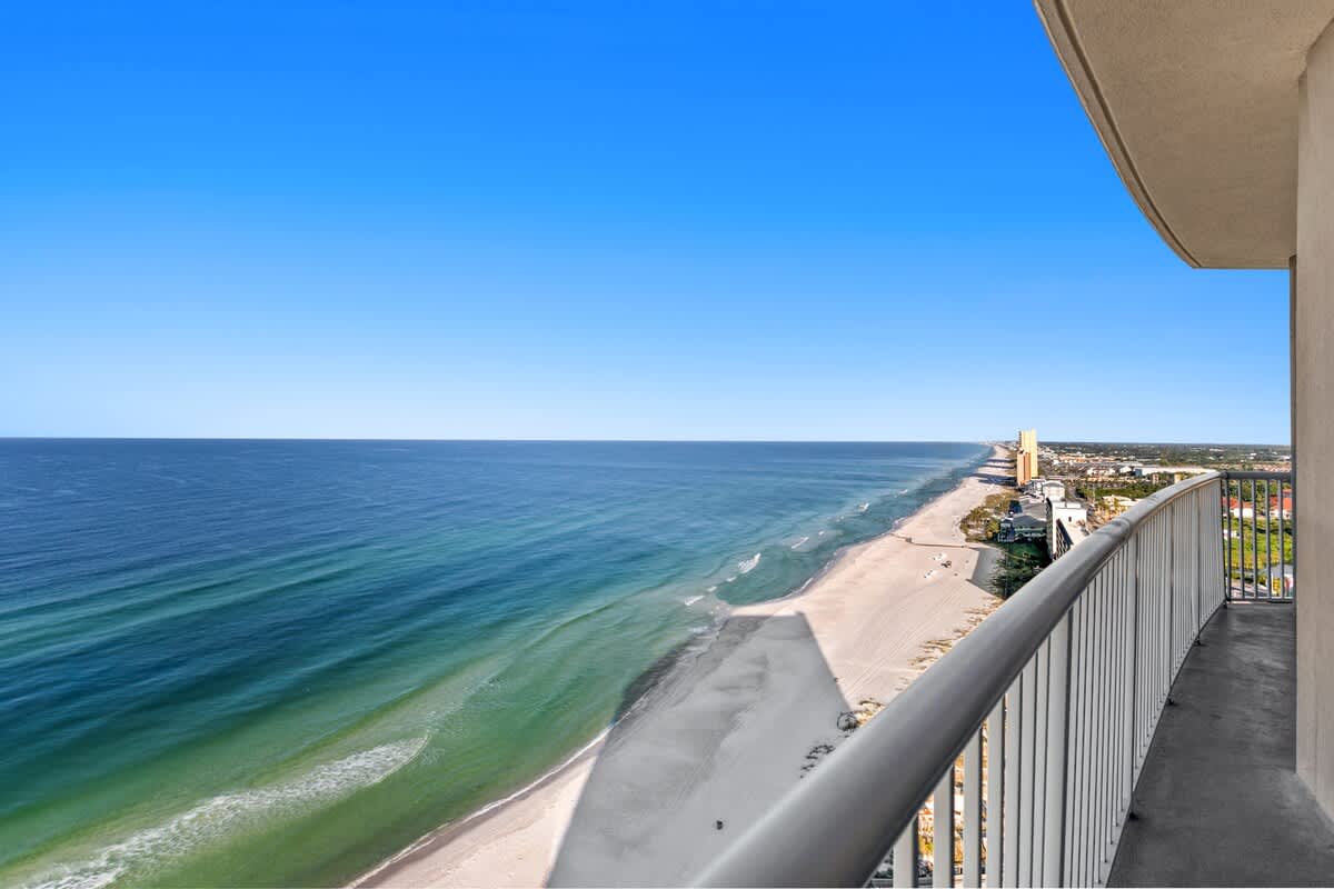 Curved balcony offering a sweeping view of a long, sandy beach and the turquoise ocean stretching to the horizon. The coastline features distant buildings under a bright blue sky.