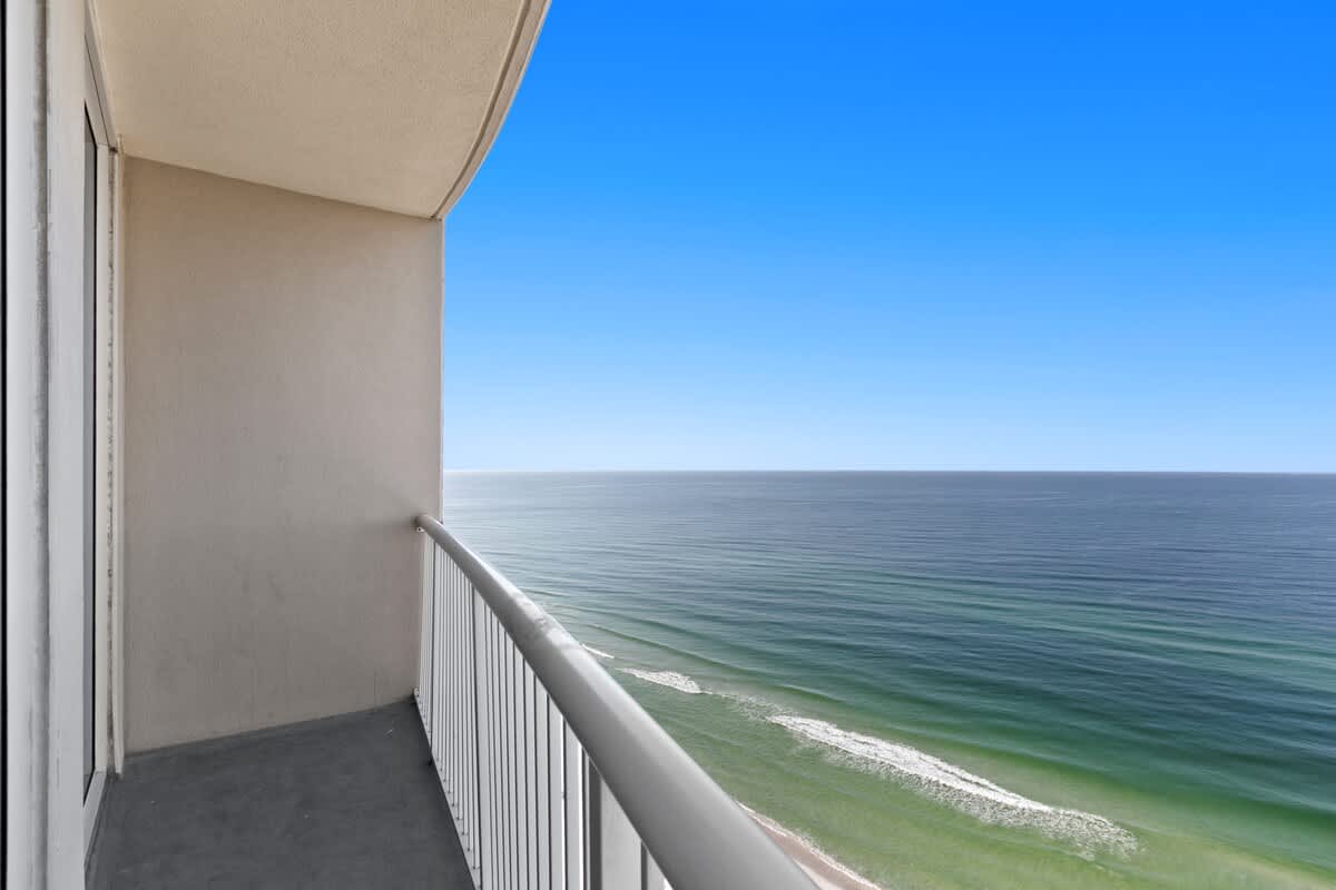 Corner view from a balcony showing a vast expanse of the blue-green ocean meeting the horizon under a clear sky. A simple metal railing runs along the edge of the concrete balcony.