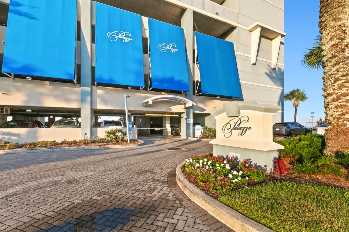 Entrance to the Palazzo condominium building, featuring a paved driveway, lush landscaping with flowers and palm trees, and a prominent sign with the property name. Blue awnings cover the parking garage entrance.