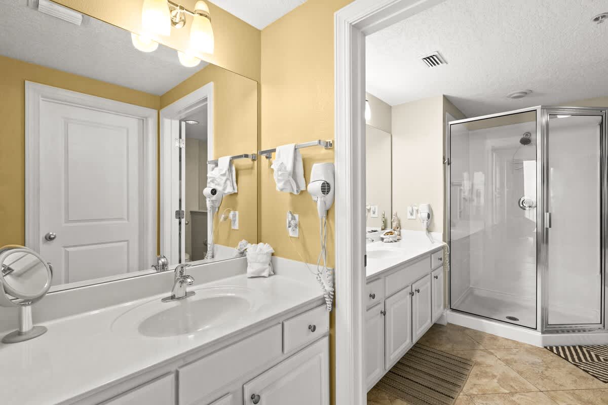 This bathroom features a single vanity with white cabinets and countertop, a large mirror, and yellow walls, with a separate shower enclosure visible and tiled flooring.