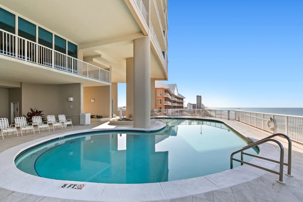 An outdoor communal pool with a curved shape and shallow entry is surrounded by light-colored tiled flooring and white lounge chairs. The pool is located on a lower level of the building, with the ocean visible in the background and other buildings in the distance.