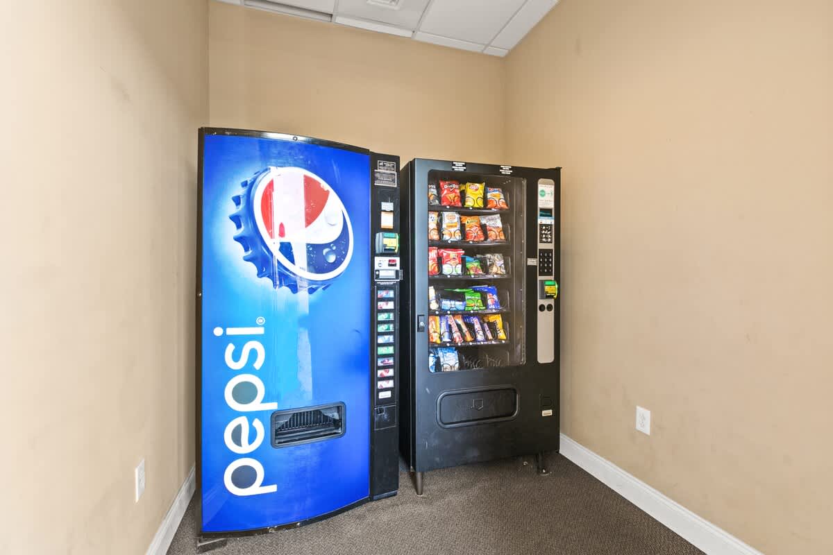 A small, carpeted alcove featuring a large blue Pepsi soda vending machine and a black snack vending machine against light beige walls. This area provides convenient access to refreshments within the property.