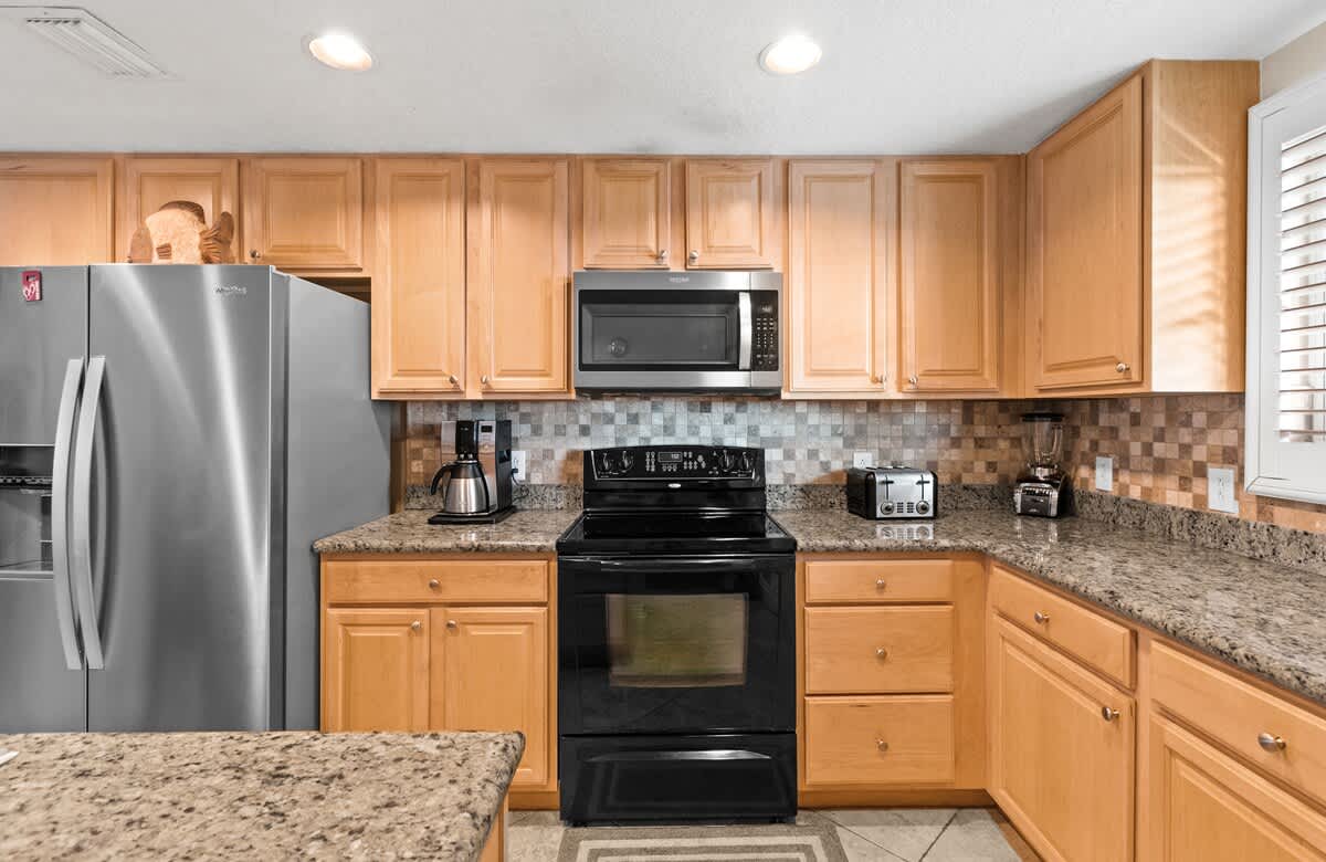 The kitchen showcases light wood cabinetry, granite countertops, and a mosaic tile backsplash, equipped with a black electric range, microwave, and stainless steel refrigerator. Various small appliances are visible on the counter.