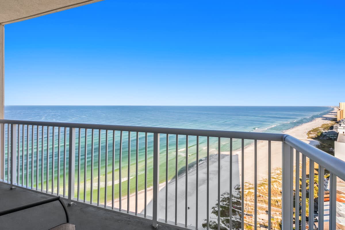 High-angle view from a balcony overlooking a wide sandy beach and the vast blue-green ocean with gentle waves. A metal railing frames the foreground, and the horizon is clear under a bright sky.