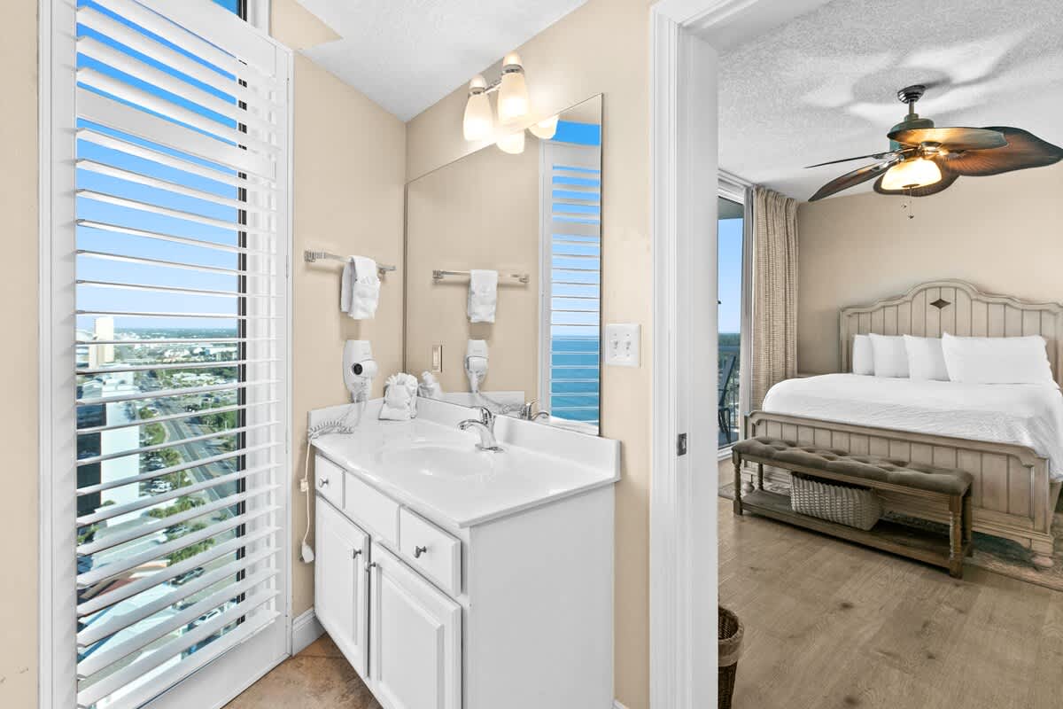 Bathroom with a white vanity, single sink, and a large mirror, featuring a window with louvered shutters that reveal an ocean view. An open doorway connects to a bedroom with a white bed and ceiling fan.