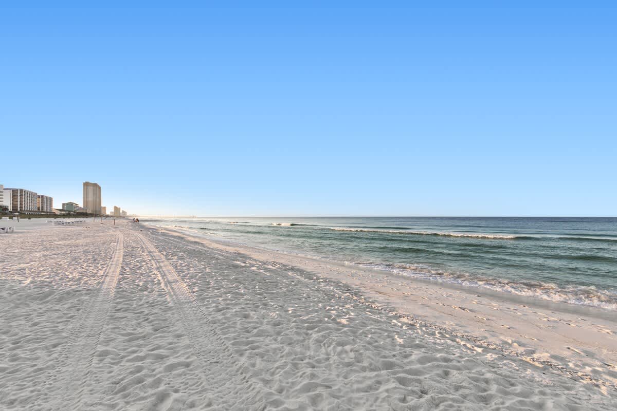 Wide shot of a pristine sandy beach with gentle waves rolling onto the shore, under a clear blue sky. Tall condominium buildings line the distant coastline, suggesting a vibrant resort area.