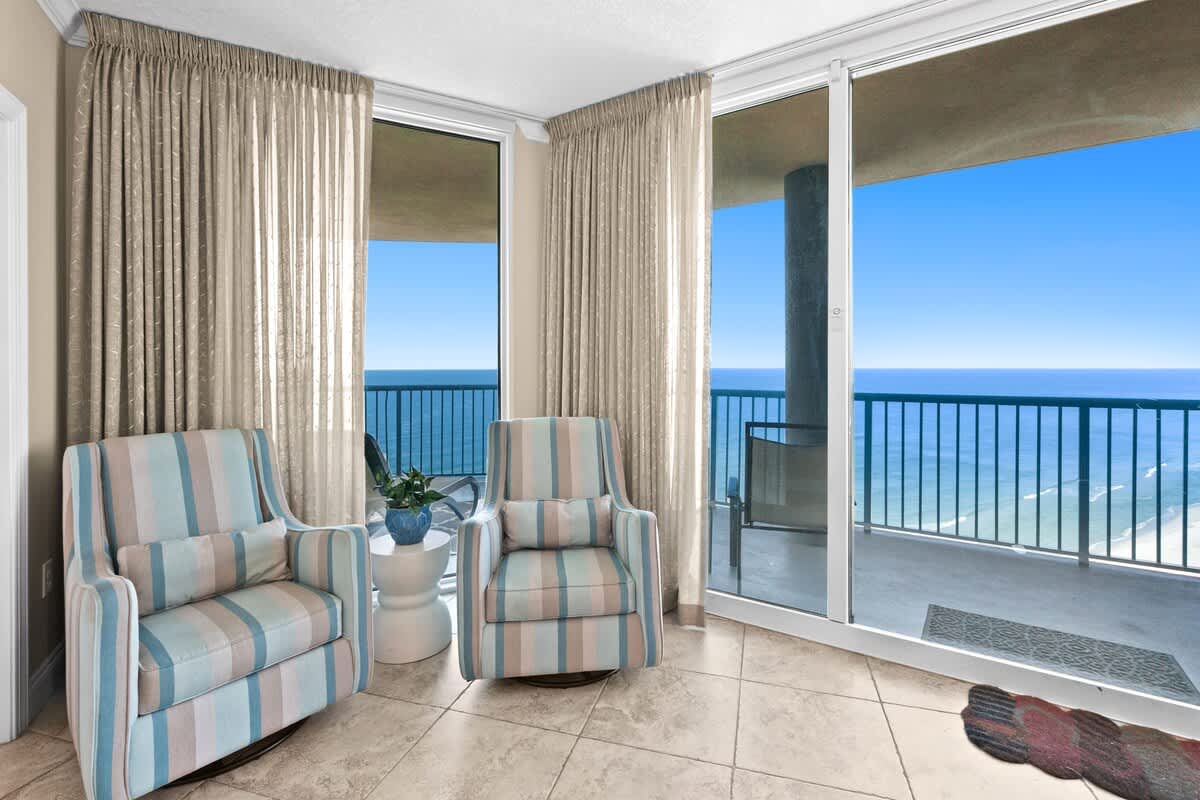 A bright seating nook features two comfortable striped armchairs and a small white side table, positioned directly in front of expansive floor-to-ceiling windows offering uninterrupted ocean views. The room has light tile flooring.