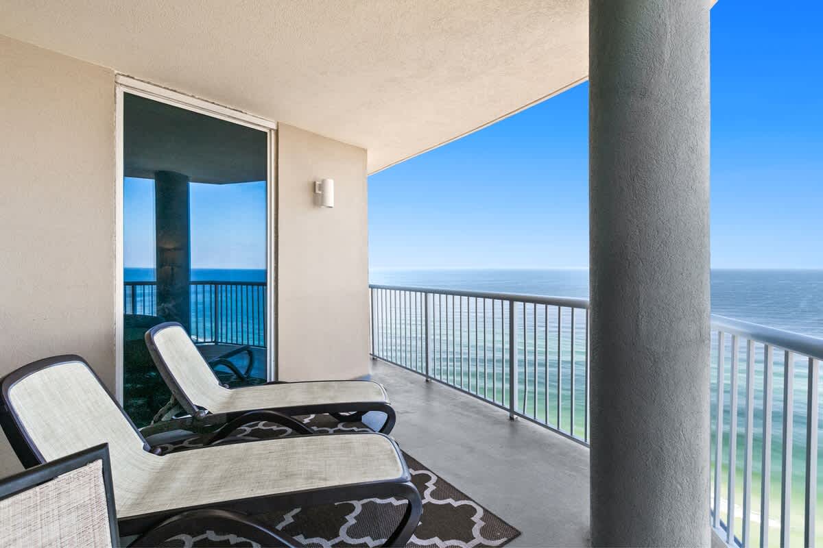 Expansive outdoor balcony featuring two lounge chairs and an outdoor rug, supported by concrete pillars and secured with metal railings, providing an unobstructed view of the turquoise ocean and white sand beach below.