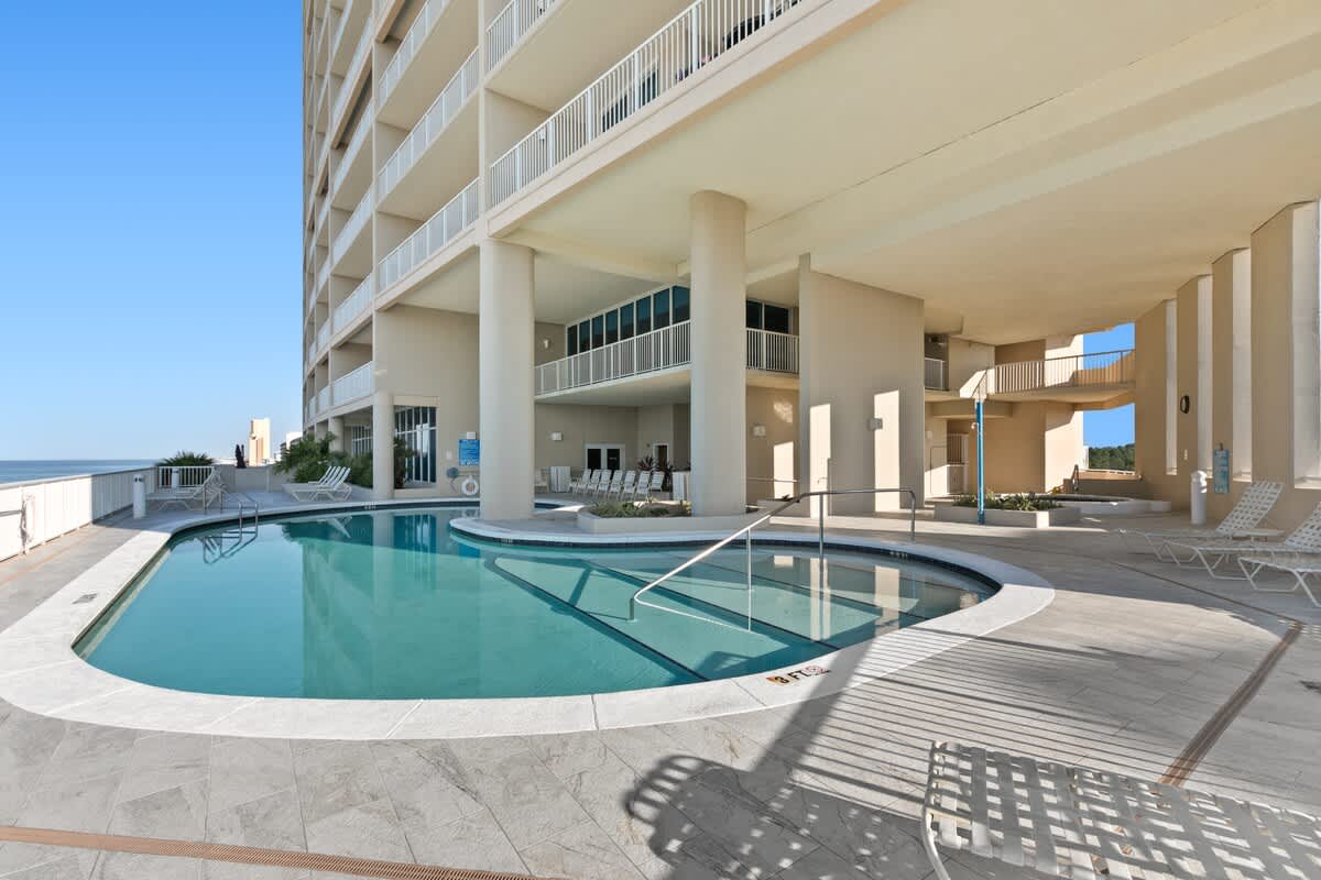 A large outdoor communal pool area with a curved pool and shallow entry is surrounded by light-colored tiled flooring. The pool is situated beneath the multi-story building, with the ocean visible in the distance and lounge chairs nearby.