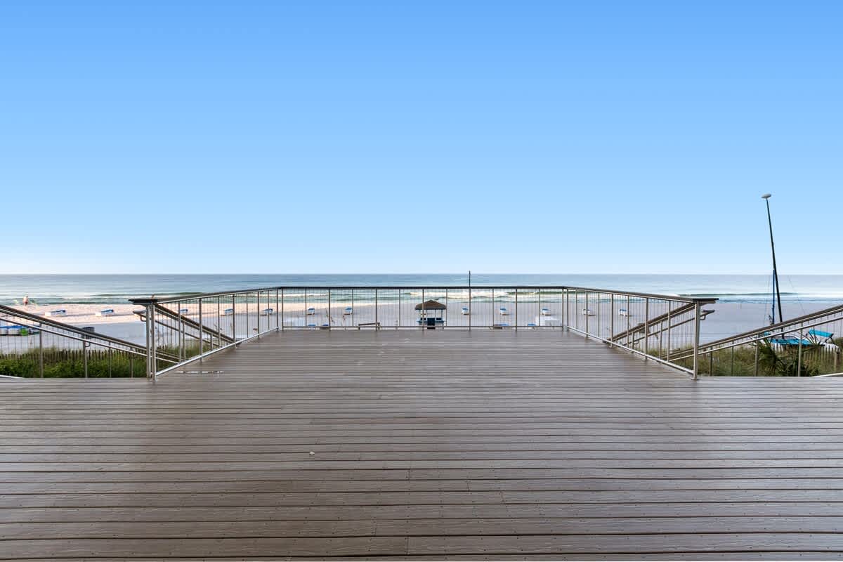 Expansive wooden deck with sturdy metal railings, providing direct and scenic access to the sandy beach and ocean beyond. A small beach hut is visible in the distance on the sand.