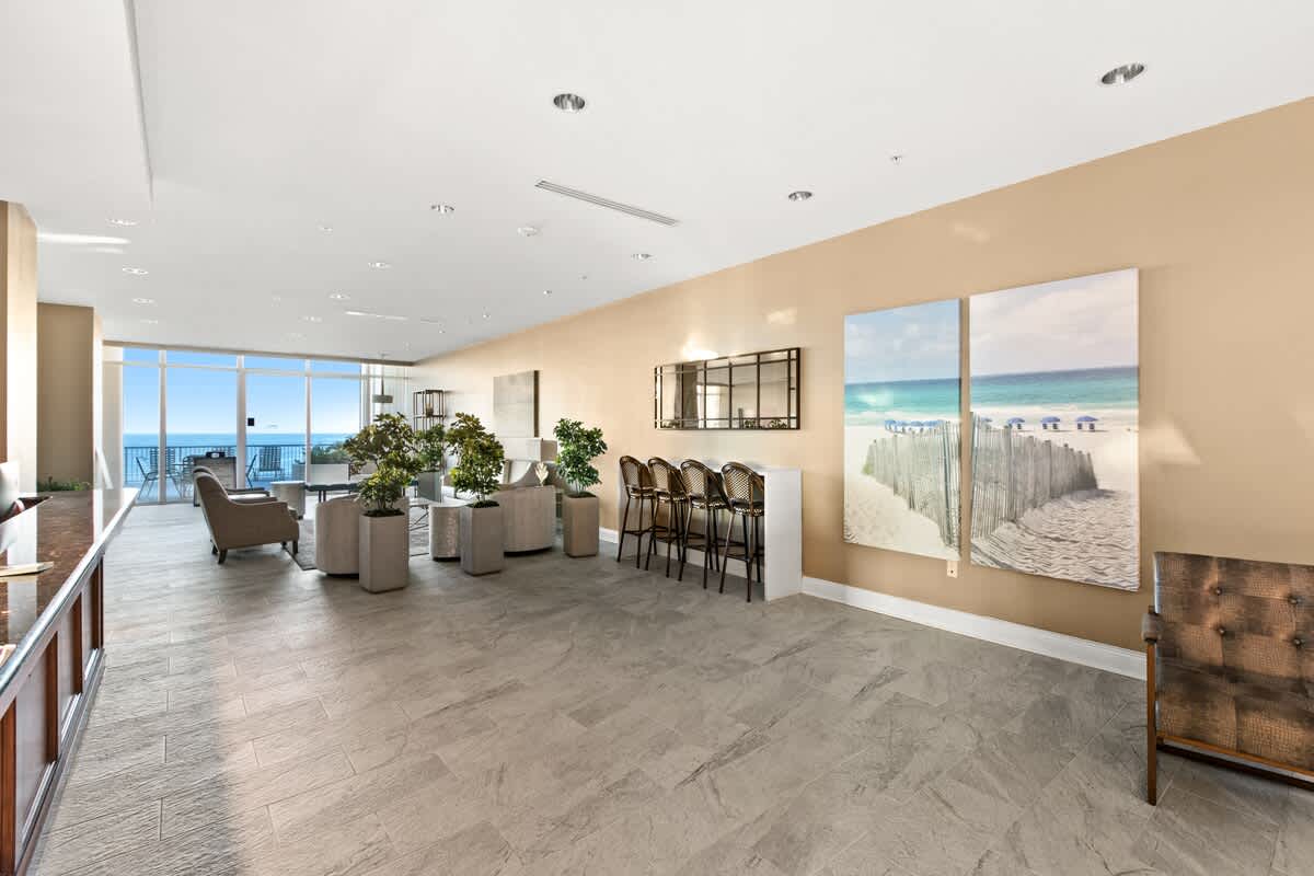 Modern and airy lounge area within the lobby, featuring light-colored tiled floors and contemporary decor with potted plants. Large windows provide panoramic ocean views, and beach-themed artwork adorns the walls.