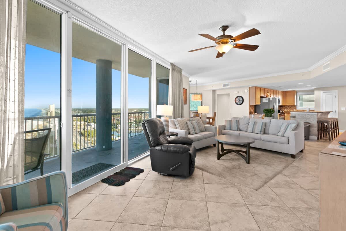 The living room features light-colored sofas, a dark leather recliner, and a round coffee table on a rug, with large sliding glass doors leading to a balcony offering views of the ocean and coastline. Light tile flooring extends throughout the space.