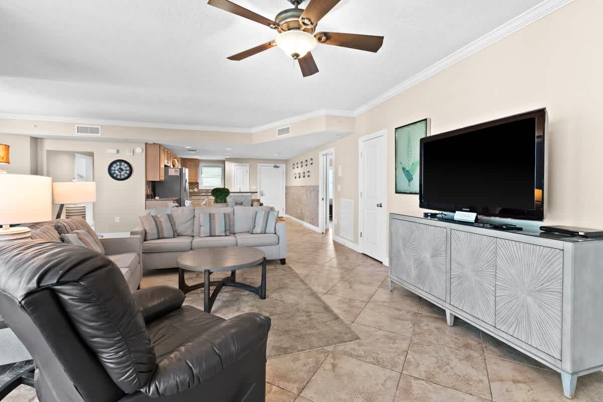 A cozy living room features a large flat-screen TV on a light-colored console, flanked by two light sofas and a dark leather recliner, with a hallway visible in the background. The room has light tile flooring and a ceiling fan.