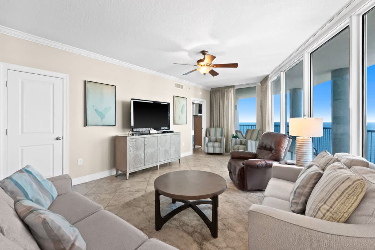 The living room is furnished with two light-colored sofas, a dark leather recliner, and a round coffee table on a rug, facing a large flat-screen TV on a light console. Large sliding glass doors provide access to a balcony with ocean views.