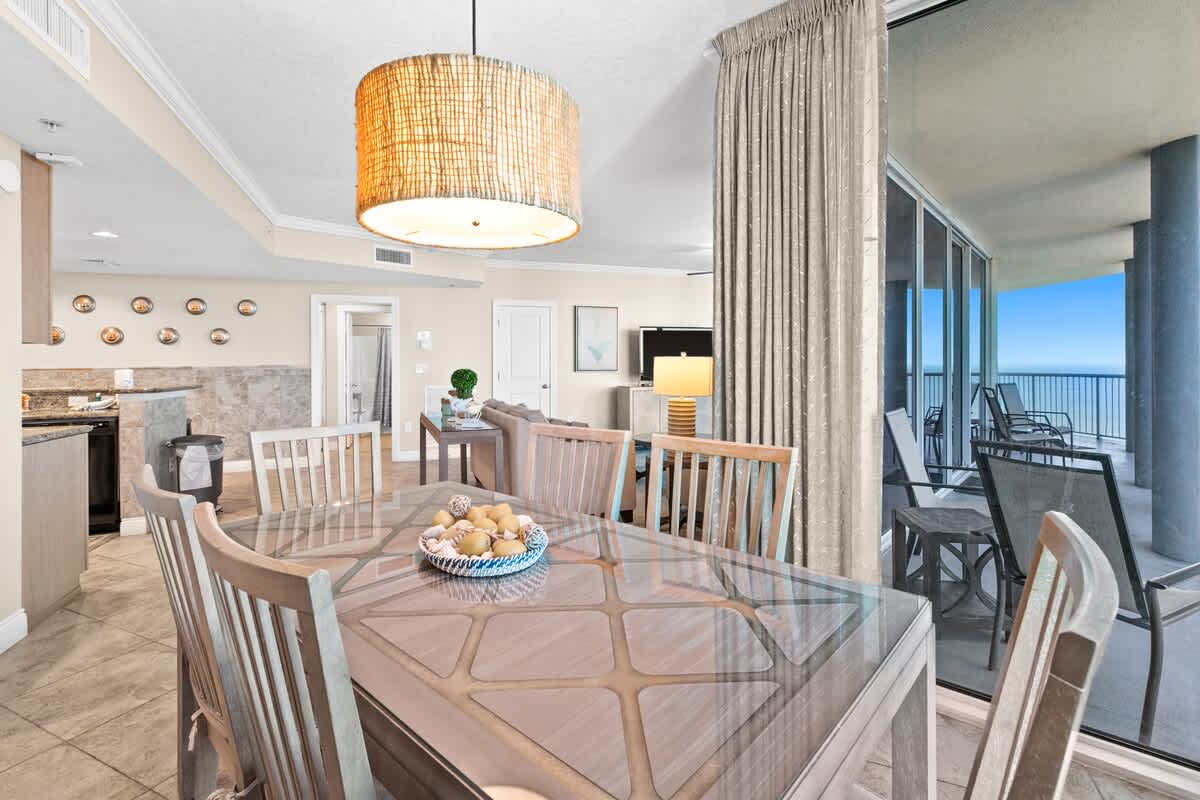 The dining area, with its glass-top table and wooden chairs, is adjacent to the kitchen bar and offers access to a balcony through sliding glass doors, with a glimpse of the living room beyond. A woven pendant light hangs above the table.