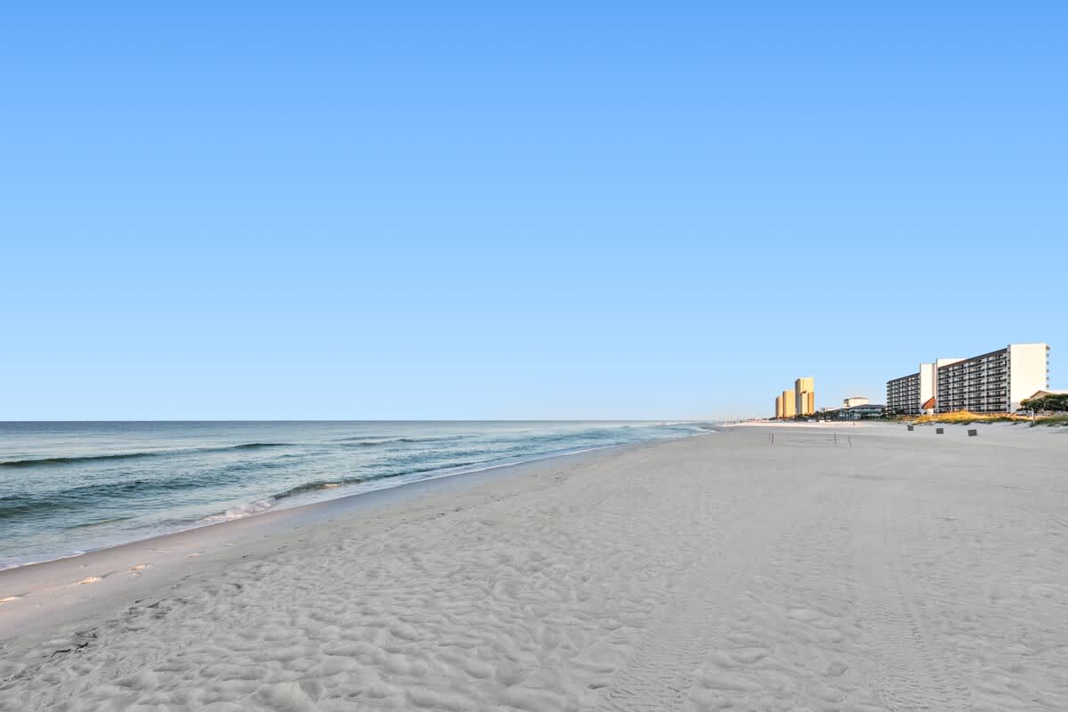 Expansive view of a pristine white sand beach stretching along the coastline, with the turquoise ocean gently lapping the shore. Several multi-story buildings are visible in the distance under a bright blue sky.