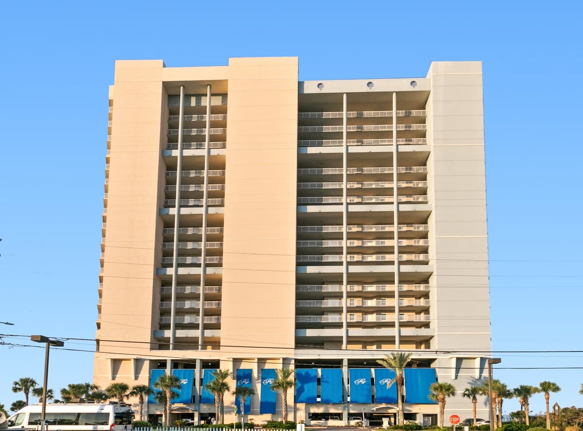 Tall, modern condominium building with light beige and gray facades, featuring numerous balconies. The ground level shows a parking garage entrance with large blue awnings displaying the "Palazzo" logo.