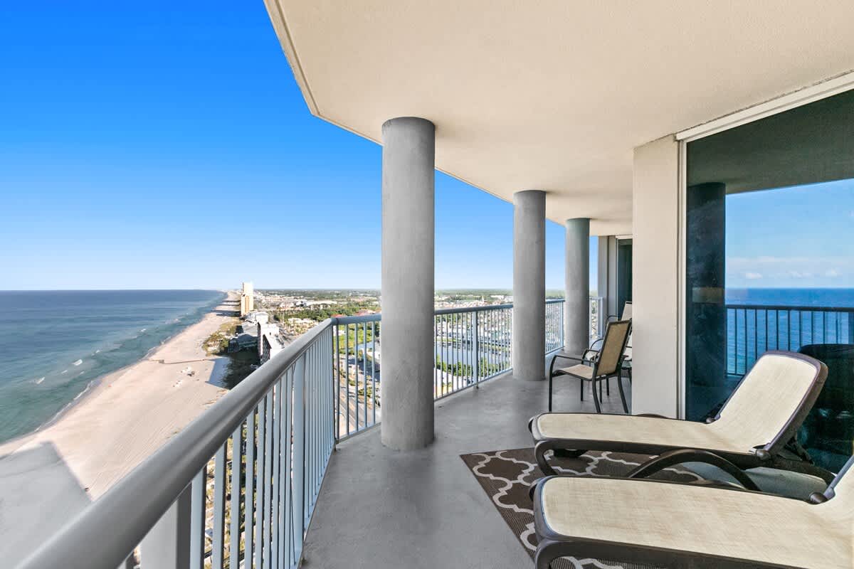Long, curved concrete balcony with multiple pillars and a metal railing, offering expansive views of the coastline, ocean, and city. Outdoor lounge chairs and a rug are placed on the balcony.