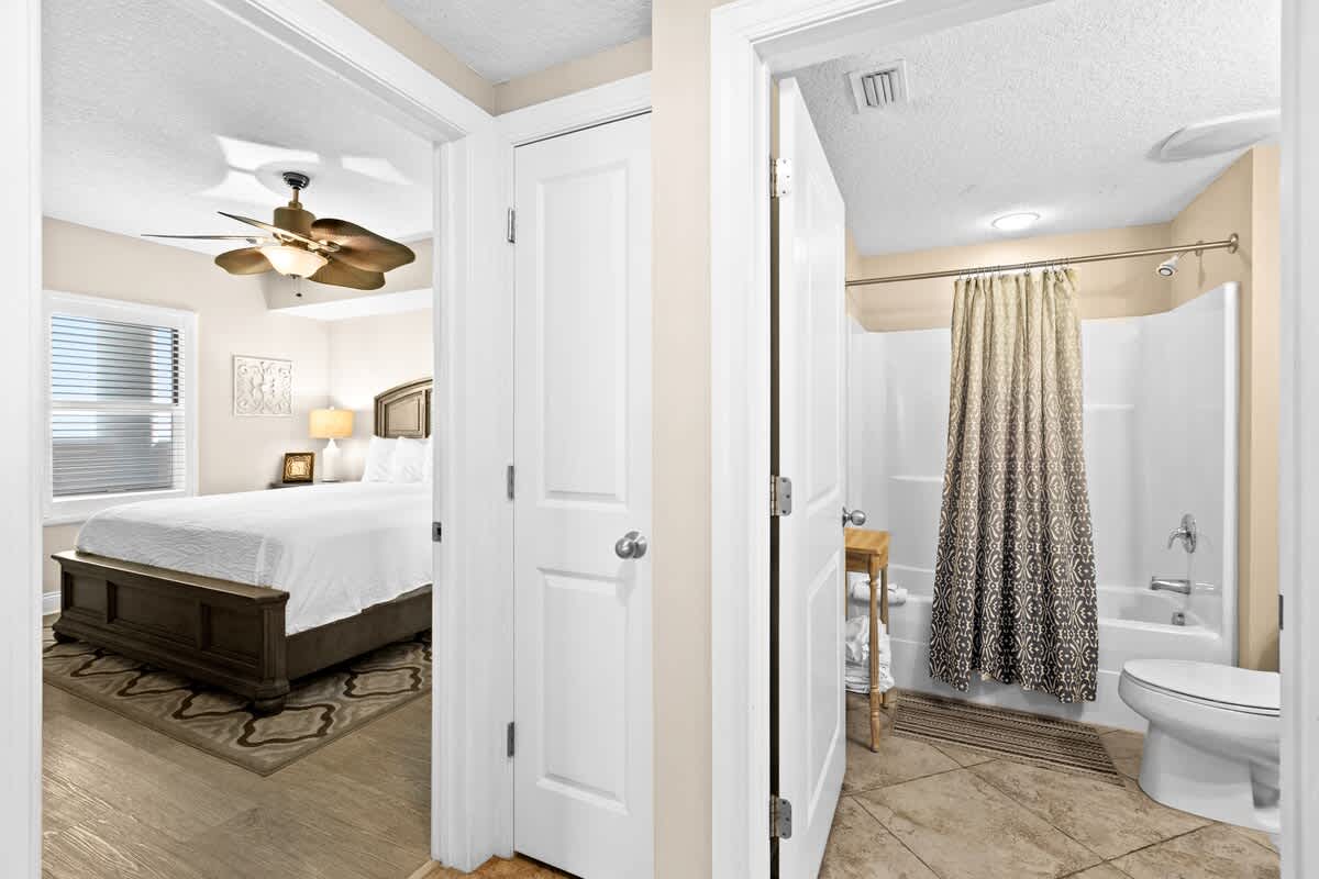 A hallway with light wood flooring leads to an open doorway revealing a bedroom with a bed and a separate doorway to a bathroom with a shower/tub combo.