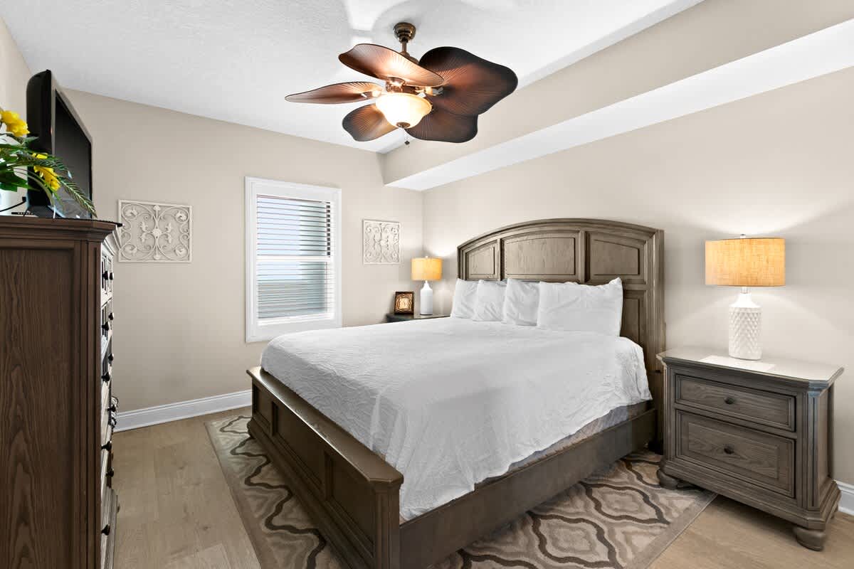 Cozy bedroom featuring a wooden king-sized bed with white bedding, light wood-look flooring, and a decorative ceiling fan. Two matching nightstands with lamps flank the bed, and a tall dresser is visible on the left.