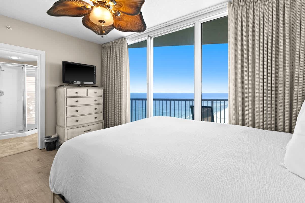 Bedroom with a white bed, light wood-look flooring, a white dresser topped with a TV, and a tropical leaf-bladed ceiling fan. Large sliding glass doors open to a balcony with stunning ocean views.