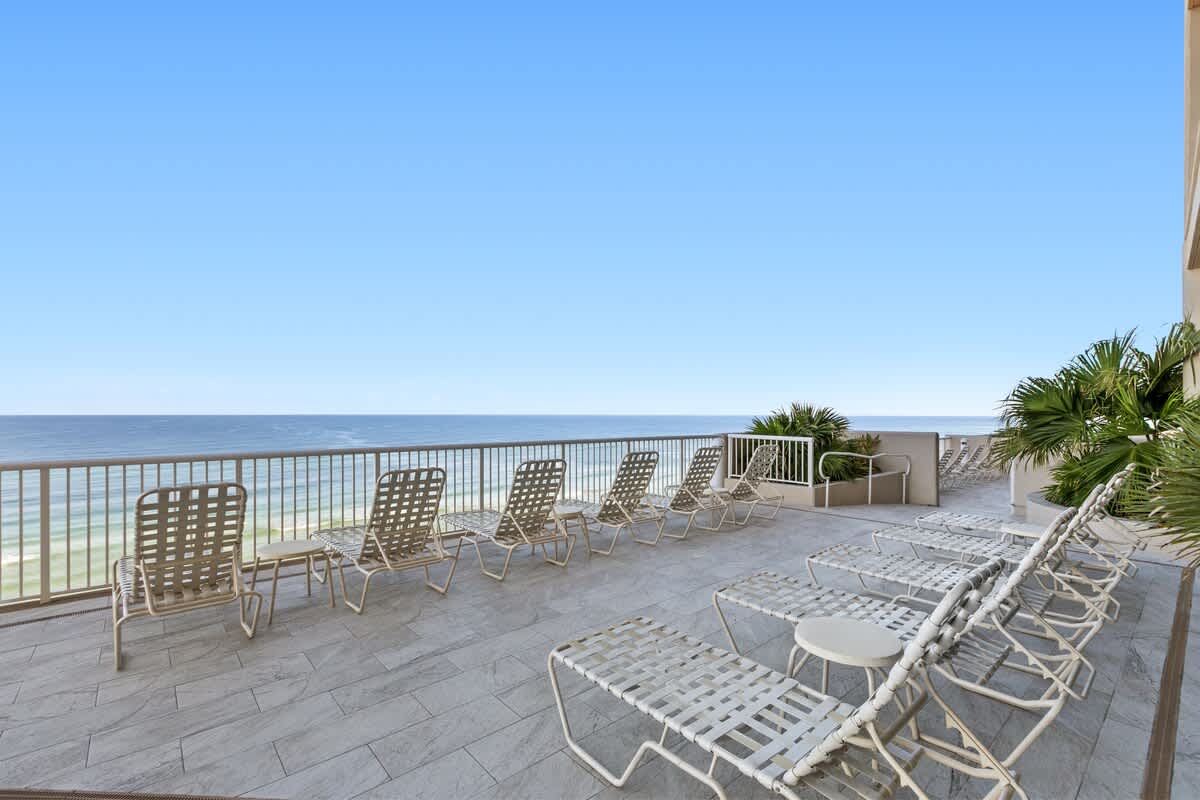 A spacious outdoor deck with light-colored tiled flooring is furnished with numerous white mesh lounge chairs. The deck offers panoramic views of the ocean and sandy beach below, framed by a white railing and some lush greenery.