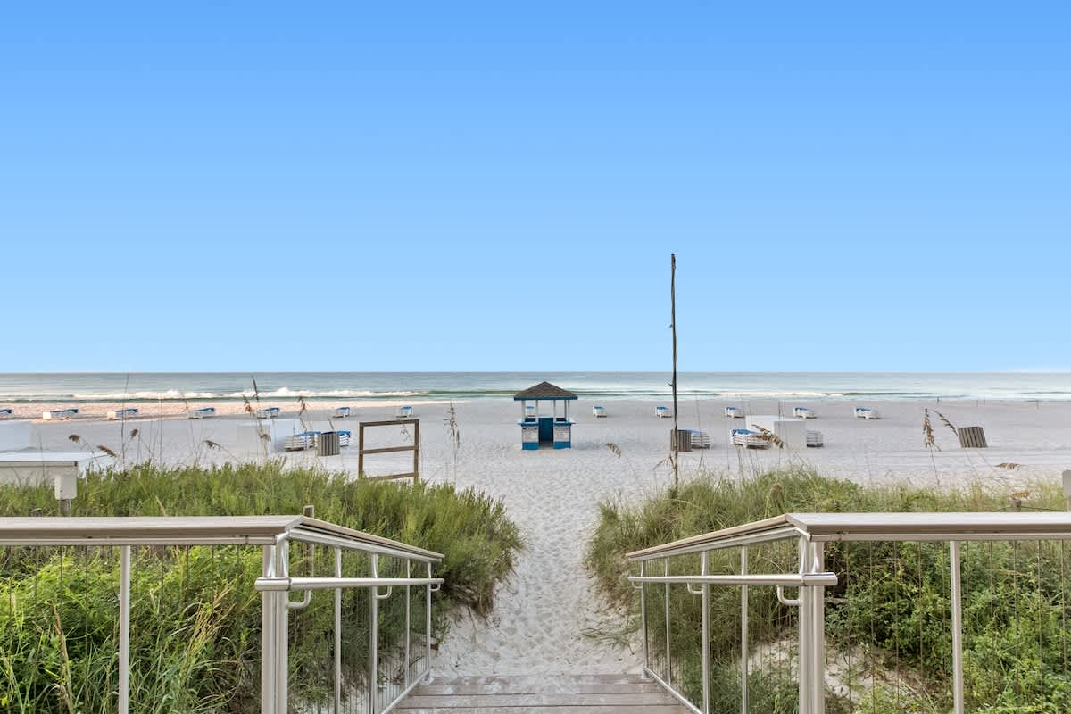 Wooden walkway and stairs leading down through green dunes to a wide, pristine sandy beach with beach chairs and umbrellas. The calm blue ocean stretches to the horizon under a clear sky.