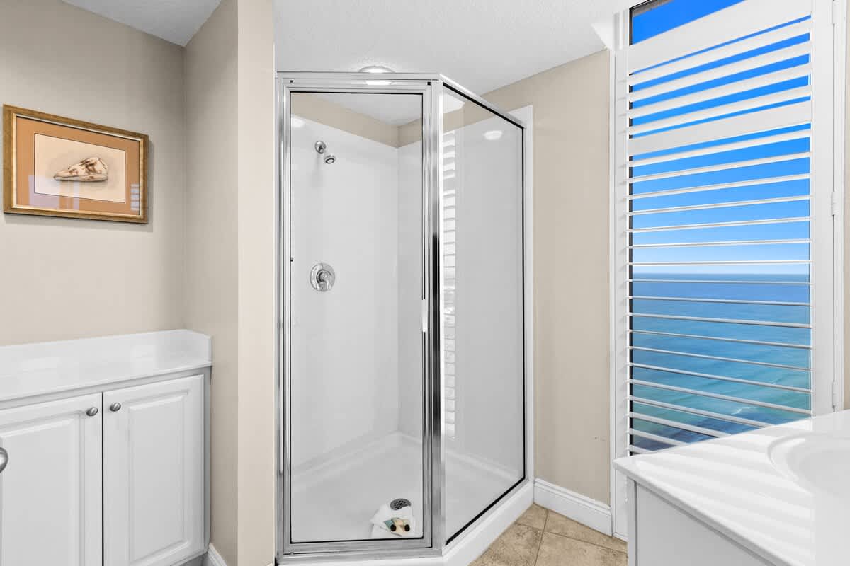 Modern bathroom featuring a corner glass shower, a white vanity with a single sink, and a large window with white louvered shutters providing a partial ocean view.
