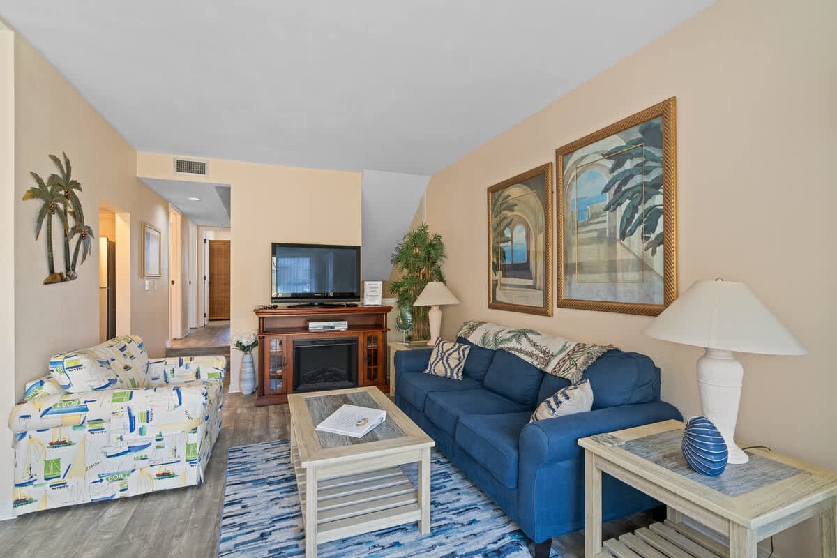 The living room features a blue sofa, a patterned armchair, a TV stand with an electric fireplace, and a light wood coffee table, with a hallway visible to the left.
